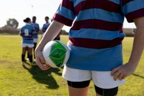 Vista frontal de la sección central de una joven jugadora de rugby caucásica adulta parada en un campo de rugby sosteniendo una pelota de rugby, con sus compañeras de equipo hablando juntas en el fondo - foto de stock