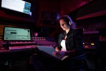 Вид спереди на молодую кавказскую звукоинженершу, сидящую и работающую за микшерским столом в студии звукозаписи с помощью ноутбука и в наушниках — стоковое фото