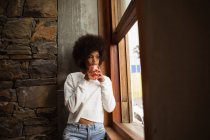 Nahaufnahme einer jungen Frau mit gemischter Rasse, die am Fenster steht, hinausblickt und eine Tasse Kaffee in der Hand hält — Stockfoto