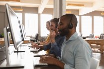 Side view close up of a young African American man and a young Caucasian woman and man sitting at desks using computers and talking on phone headsets in a creative office — Stock Photo