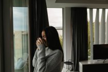Side view of a young Caucasian brunette woman wearing a grey sweater, standing by a window drinking a cup of coffee with eyes closed — Stock Photo