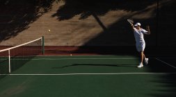 Вид сбоку на молодого кавказца, играющего в теннис в солнечный день, бьющего по мячу со стеной позади него — стоковое фото