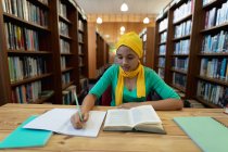 Вид спереди на молодую азиатскую студентку в хиджабе, которая делает заметки и учится в библиотеке — стоковое фото
