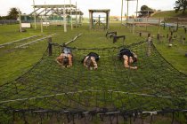 Vue de face de deux jeunes femmes caucasiennes et d'un jeune homme caucasien rampant sous un filet dans un gymnase extérieur lors d'une séance d'entraînement de bootcamp — Photo de stock