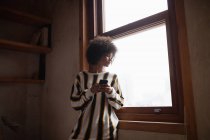 Vista frontal de una joven mujer de raza mixta de pie y mirando por una ventana en casa, utilizando un teléfono inteligente - foto de stock