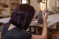 Vista posteriore da vicino di una giovane ceramista caucasica inginocchiata e che lavora su una fiaschetta di argilla su un tavolo usando uno strumento di modellazione — Foto stock