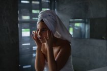 Vista frontal close-up de uma jovem mulher morena caucasiana vestindo uma toalha de banho e com o cabelo envolto em uma toalha, olhando no espelho e massageando o rosto com os dedos em um banheiro moderno — Fotografia de Stock