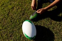 Sección baja de alto ángulo de la jugadora de rugby hembra arrodillada y atando su bota en un campo de rugby, con la pelota a su lado - foto de stock