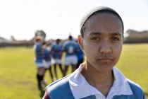 Porträt einer jungen erwachsenen gemischten Rugbyspielerin, die auf einem Rugbyfeld steht und in die Kamera blickt, während ihre Teamkollegen im Hintergrund miteinander reden — Stockfoto