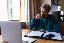 Вид спереди пожилой белой женщины, сидящей за столом дома и пьющей кофе со смартфоном и ноутбуком перед ней — стоковое фото