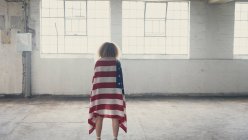 Вид сзади на молодую кавказскую женщину с вьющимися волосами и американским флагом за плечами, стоящую в пустом складе — стоковое фото