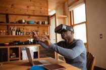 Vue de côté gros plan d'un jeune Afro-Américain portant un casque VR assis à la table de la cuisine à la maison avec un ordinateur portable, les bras levés et les mains tendues — Photo de stock