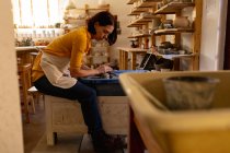 Vue latérale d'une jeune potière caucasienne assise et travaillant avec de l'argile sur une roue de potiers dans un atelier de poterie, avec équipement au premier plan — Photo de stock