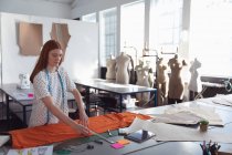 Vue latérale d'une jeune étudiante de mode caucasienne travaillant sur un design mesurant le tissu orange dans un studio au collège de mode, avec des mannequins en arrière-plan — Photo de stock