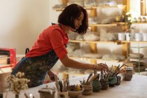 Вид сбоку молодой кавказской женщины-гончара, стоящей и выбирающей инструмент из горшков с инструментами на рабочем столе в мастерской керамики — стоковое фото