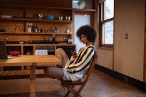 Вид сбоку на молодую женщину смешанной расы с помощью смартфона, сидящую дома за кухонным столом — стоковое фото