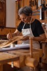 Seitenansicht einer älteren kaukasischen Geigenbauerin, die auf einer Werkbank in ihrer Werkstatt am Körper einer Geige arbeitet, im Hintergrund hängen Werkzeuge an der Wand — Stockfoto