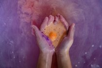 Acercamiento de las manos ahuecadas de una mujer en un baño que contiene sales de baño naranjas efervescentes en el agua del baño rosa - foto de stock
