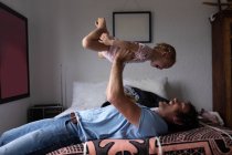 Side view of a young Caucasian father holding his baby above him and lying on a bed — Stock Photo