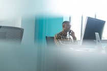 Вид сбоку на молодого афроамериканца, сидящего за столом за компьютером и разговаривающего на смартфоне в современном офисе креативного бизнеса, с размытым передним план — стоковое фото