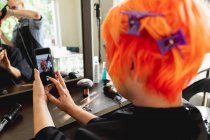 Rear view close up of a middle aged Caucasian male hairdresser and a young Caucasian woman taking a selfie while having her hair colored bright red in a hair salon, reflected in a mirror — Stock Photo