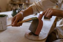 Primer plano de las manos de una alfarera pintando un esmalte de color sobre un matraz de arcilla en una mesa de trabajo en un estudio de cerámica - foto de stock