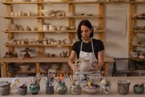 Vue de face d'une jeune potière caucasienne debout à une table de travail tenant une cruche sur une roue de baguage dans un atelier de poterie, avec des pots et des outils au premier plan — Photo de stock