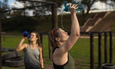 Side view of a young Caucasian woman pouring water from a bottle over her head to cool down at an outdoor gym during a bootcamp training session, another woman is drinking water in the background — Stock Photo