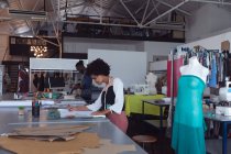 Side view of a young mixed race female fashion student working on a design in a studio at fashion college, with a mannequin withe dress on in the foreground and a male student working in the background — Stock Photo