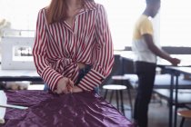 Передній вид середньої секції молодої кавказької студентки мод, що вирізає фіолетову тканину під час роботи над дизайном у студії в коледжі моди, з студентом, який працює на фоні — стокове фото