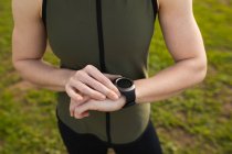 Frontansicht mittlere Sektion einer Frau, die während eines Bootcamp-Trainings in einem Outdoor-Fitnessstudio steht und ihre Uhr überprüft — Stockfoto