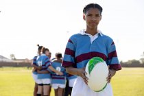 Portrait of a young adult mixed race female rugby player standing on a rugby pitch holding a rugby ball in her hands looking to camera, with her teammates talking together in the background — Stock Photo