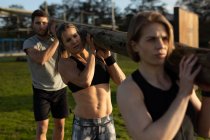 Front view of two young Caucasian women and a young Caucasian man carrying a log of wood together on their shoulders at an outdoor gym during a bootcamp training session — Stock Photo