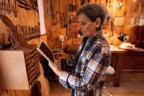 Vue latérale d'un luthier féminin caucasien aîné à l'aide d'une tablette avec des outils accrochés au mur en arrière-plan — Photo de stock