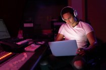 Vista frontal de perto de um jovem engenheiro de som misto sentado e trabalhando em uma mesa de mistura em um estúdio de gravação usando um laptop e usando fones de ouvido — Fotografia de Stock
