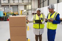 Front view close up of a young mixed race female and a young mixed race male factory workers talking in a warehouse loading bay at a factory. — Stock Photo