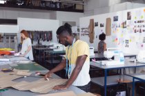 Vue latérale d'un jeune étudiant afro-américain de la mode masculine caucasienne dessinant un motif tout en travaillant sur un design dans un studio au collège de mode, avec d'autres étudiants travaillant en arrière-plan — Photo de stock