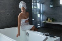 Vorderansicht einer jungen kaukasischen Frau mit Badetuch und in ein Handtuch gehüllten Haaren, die am Rand der Badewanne sitzt und in einem modernen Badezimmer das Wasser berührt — Stockfoto