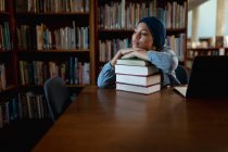 Vista frontale da vicino di una giovane studentessa asiatica che indossa un turbante appoggiata su un mucchio di libri e che studia in una biblioteca — Foto stock