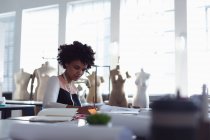 Frontansicht einer jungen Studentin mit gemischter Rasse, die an einem Tisch sitzt und in einem Atelier der Modehochschule an einem Design arbeitet — Stockfoto