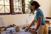 Vista laterale di una giovane ceramista caucasica che indossa un grembiule mentre lavora con pezzi di argilla su un tavolo da lavoro davanti a una finestra in uno studio di ceramica — Foto stock