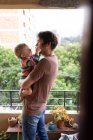Vista lateral de un joven padre caucásico sosteniendo a su bebé, de pie en un balcón con un parque en el fondo - foto de stock