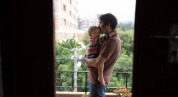 Vista lateral de un joven padre caucásico sosteniendo a su bebé, de pie en un balcón con un parque en el fondo - foto de stock