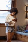 Nahaufnahme einer jungen Mischlingsfrau, die vor einem Fenster steht und zu Hause ein Smartphone benutzt — Stockfoto