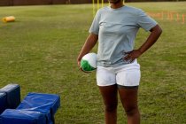 Vista frontale sezione centrale del giocatore di rugby femminile in piedi su un campo sportivo con la mano sull'anca che tiene una palla da rugby durante una sessione di allenamento — Foto stock