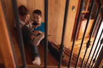 Vista frontal close-up de dois meninos pré-adolescentes caucasianos sentados em uma escada em casa, usando um smartphone — Fotografia de Stock