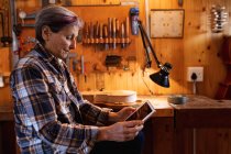Side view of a senior Caucasian female luthier using a tablet computer with tools hanging up on the wall in the background — Stock Photo