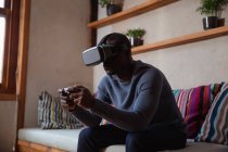 Vista lateral de perto de um jovem afro-americano vestindo um fone de ouvido VR e usando um bloco de jogo sentado em um sofá em casa — Fotografia de Stock