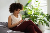 Vista lateral de cerca de una joven mujer de raza mixta sentada en una bolsa de frijoles y usando una tableta en una oficina creativa - foto de stock