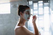 Vue de côté gros plan d'une jeune femme brune caucasienne portant une serviette de bain regardant dans le miroir et appliquant un pack visage avec une brosse dans une salle de bain moderne — Photo de stock